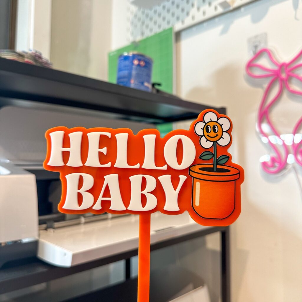 Layered acrylic cake topper featuring a flowerpot design with the phrase "Hello Baby" in a playful, layered acrylic font.