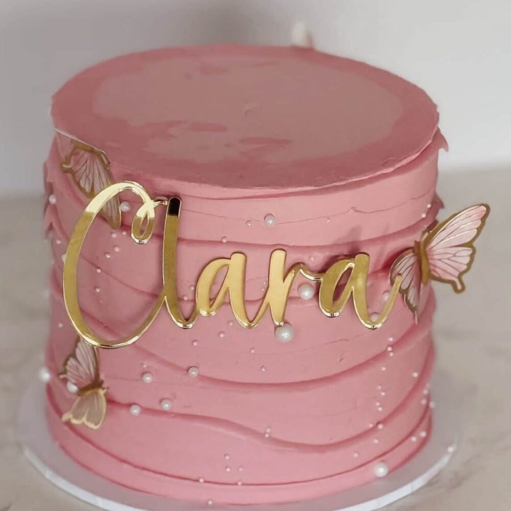 Pink buttercream cake decorated with gold “Clara” topper, pearls and butterfly accents.