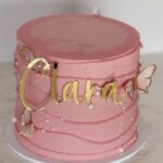 Pink buttercream cake decorated with gold “Clara” topper, pearls and butterfly accents.