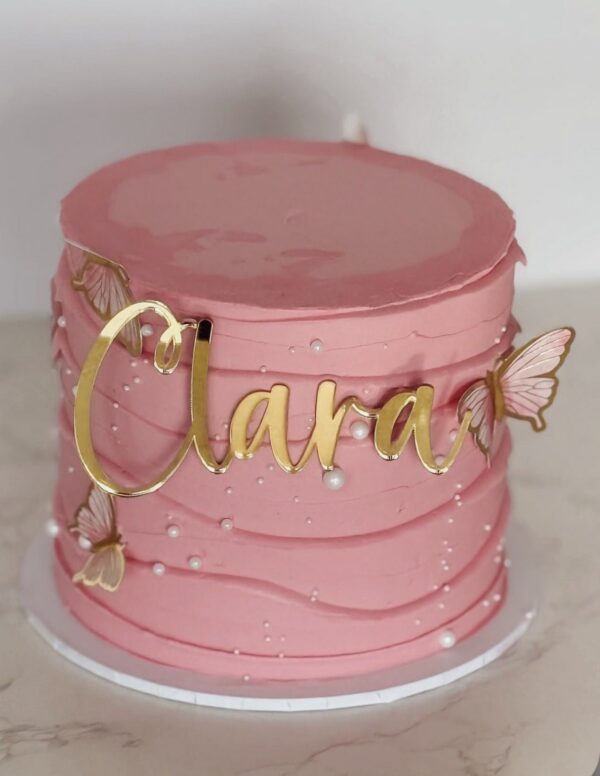 Pink buttercream cake decorated with gold “Clara” topper, pearls and butterfly accents.