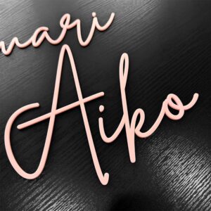 A name sign cut from a single layer of baby pink acrylic, with 'Aiko' in lighter pink cursive lettering.