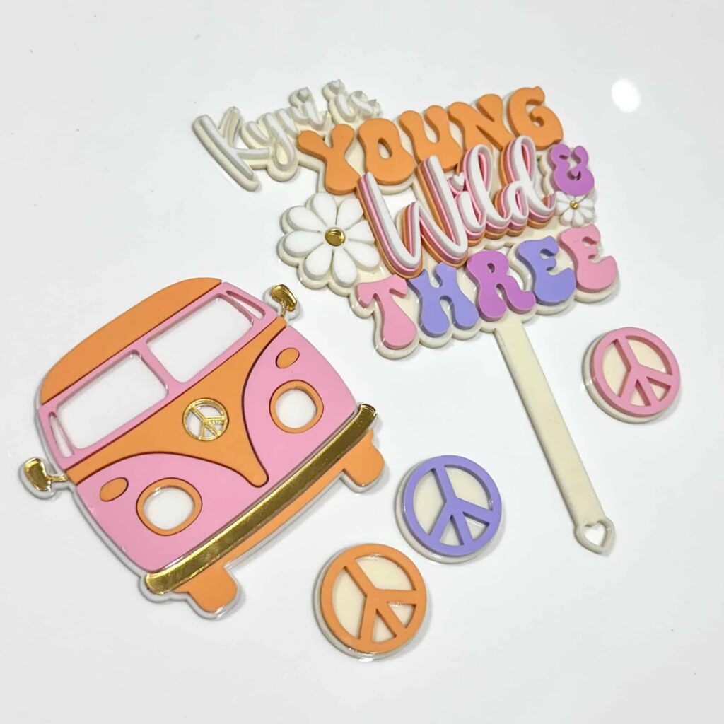 A layered acrylic cake topper with a custom design, featuring the phrase "Young, Wild & Three" in a bold, playful font.