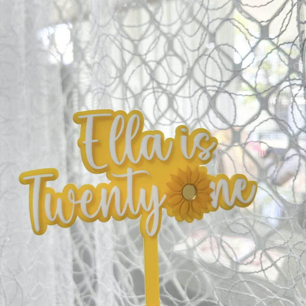 Acrylic cake topper with a sunflower design, personalised with a name and age in a bold, white font.