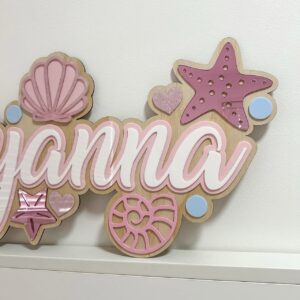Personalised mermaid name sign 'Ayyanna' with pink seashells, starfish and blue pearl accents on a wooden base.