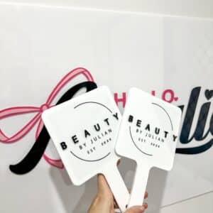 White acrylic hand fan with pink ribbon handle featuring "Beauty by Julian" branding and establishment date 2023, held in hand with partial business signage visible in background