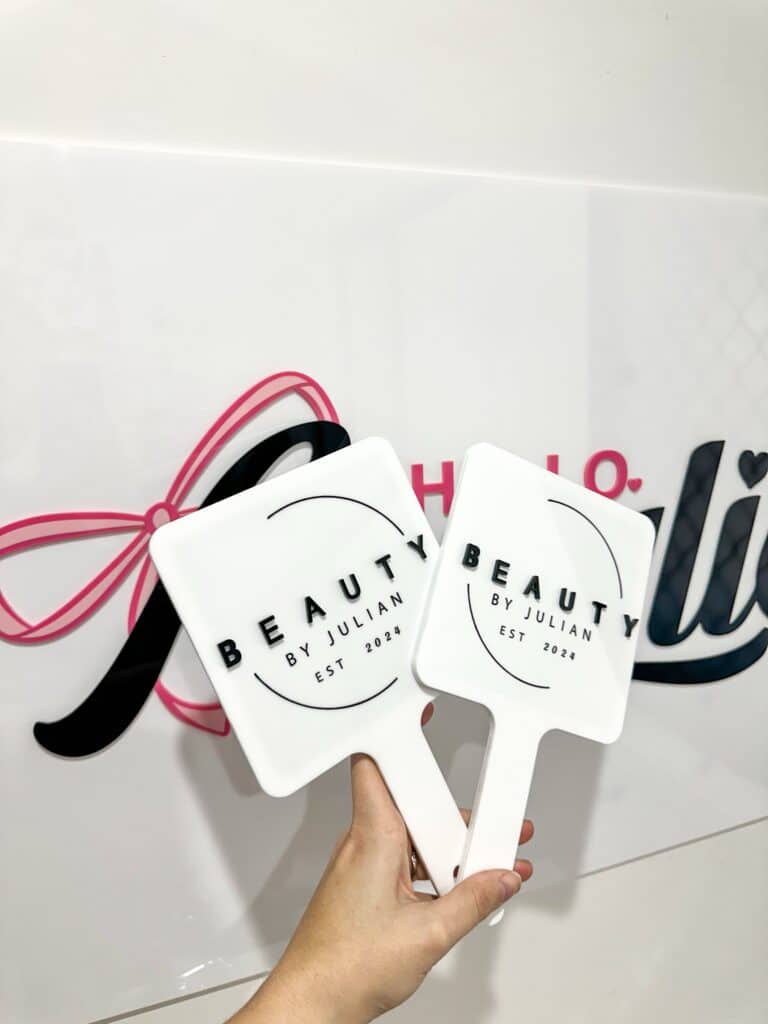 White acrylic hand fan with pink ribbon handle featuring "Beauty by Julian" branding and establishment date 2023, held in hand with partial business signage visible in background