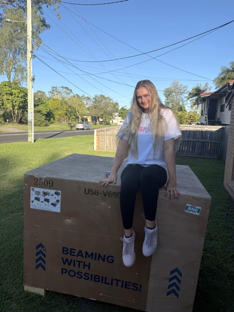 Woman sitting on a large shipping crate for a new laser engraving machine on a suburban lawn.