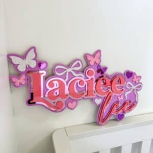 Purple and pink personalized name sign reading "Laciee" with colorful butterfly decorations, hearts, and gradient text displayed on white shelf against white wall