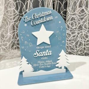Blue and white personalized Christmas countdown sign reading "The Christmas Countdown sleeps until Santa visits" with white star, evergreen trees, and custom children's names on wooden base with lace curtain background