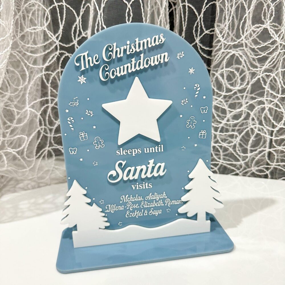 Blue and white personalized Christmas countdown sign reading "The Christmas Countdown sleeps until Santa visits" with white star, evergreen trees, and custom children's names on wooden base with lace curtain background