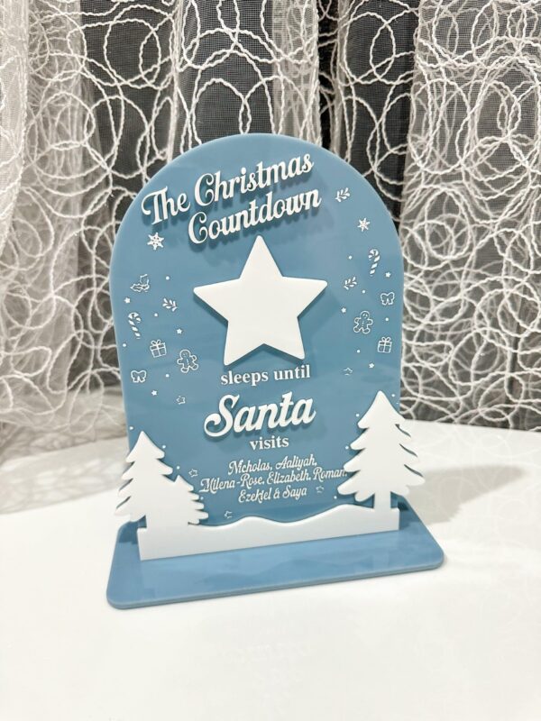 Personalized Christmas Countdown Sign - Blue Wooden Sleepover Decoration with Winter Scene Blue and white personalized Christmas countdown sign reading "The Christmas Countdown sleeps until Santa visits" with white star, evergreen trees, and custom children's names on wooden base with lace curtain background