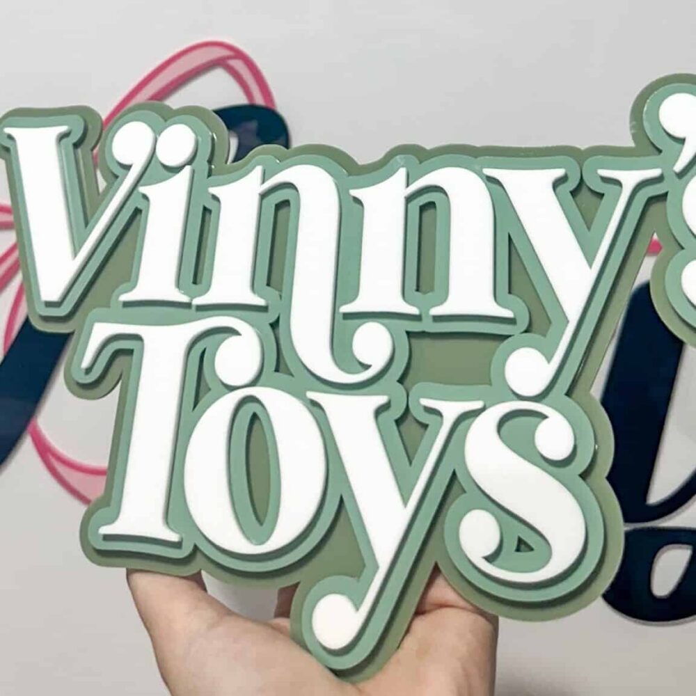A hand holding a custom layered acrylic sign that reads "Vinny's Toys" in white text with a sage green background.
