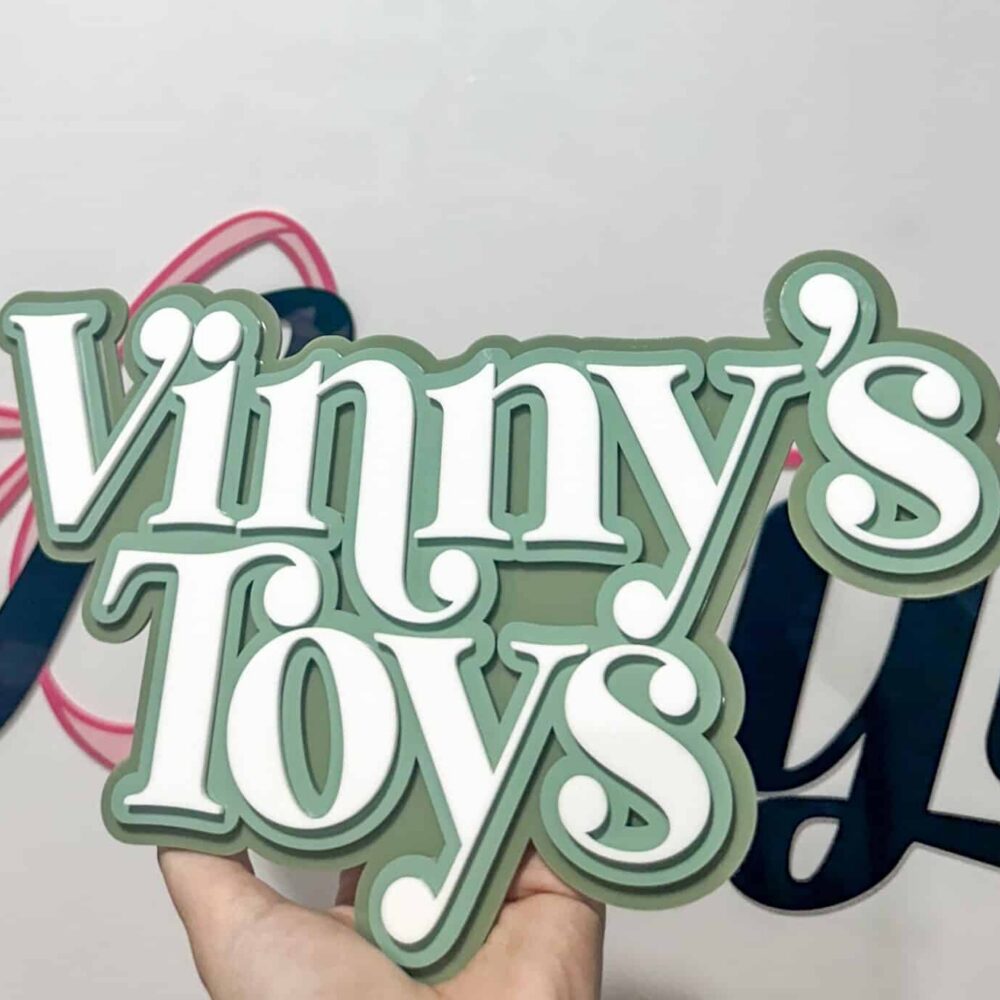 A hand holding a custom layered acrylic sign that reads "Vinny's Toys" in white text with a sage green background.