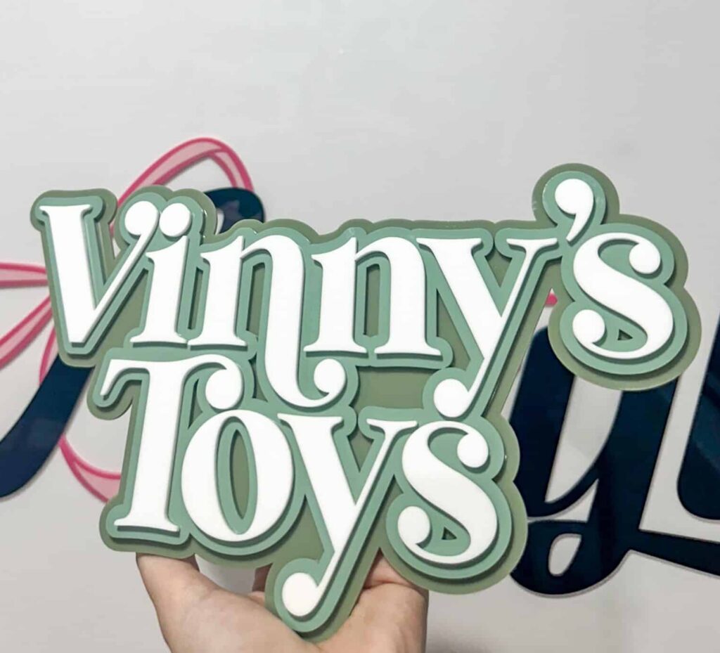 A hand holding a custom layered acrylic sign that reads "Vinny's Toys" in white text with a sage green background.