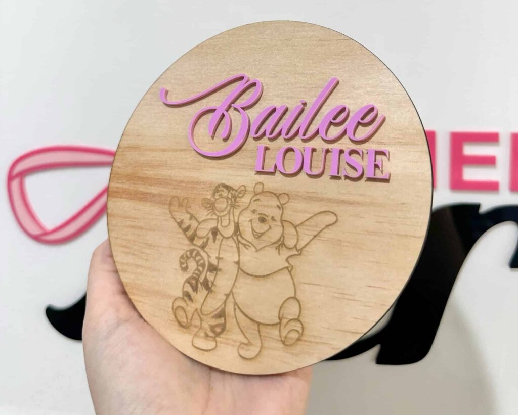 A hand holding a round wooden nursery sign with pink acrylic lettering for the name Bailee Louise and an engraved image of Winnie the Pooh and Tigger.