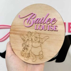 A hand holding a round wooden nursery sign with pink acrylic lettering for the name Bailee Louise and an engraved image of Winnie the Pooh and Tigger.