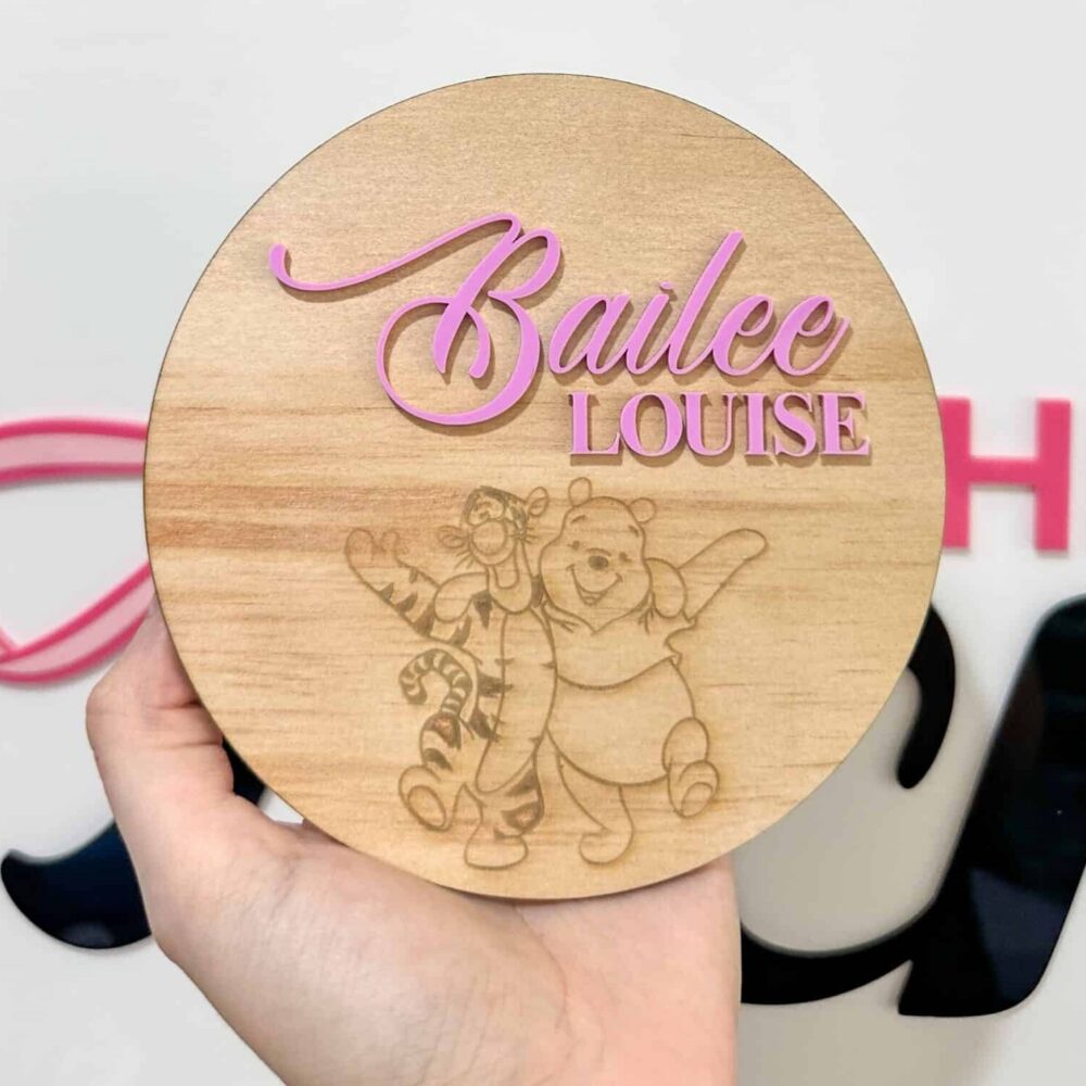 Round wooden name plaque held in hand featuring raised pink acrylic text reading "Bailee Louise" sitting above a laser-engraved line drawing of Winnie the Pooh and Tigger dancing.